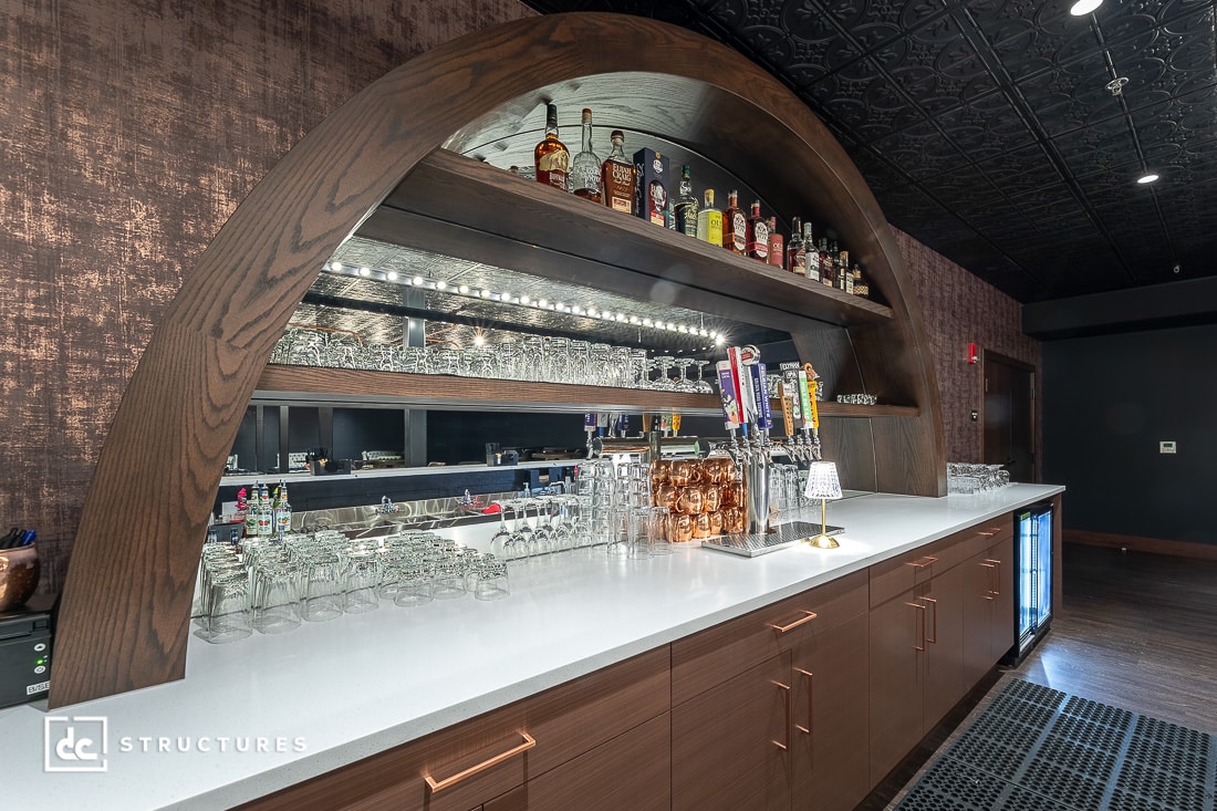 A modern bar with a white countertop, wooden arched shelving stocked with liquor bottles, glassware, beer taps, and a mirrored backsplash. Warm lighting and under-counter refrigerators are also visible.