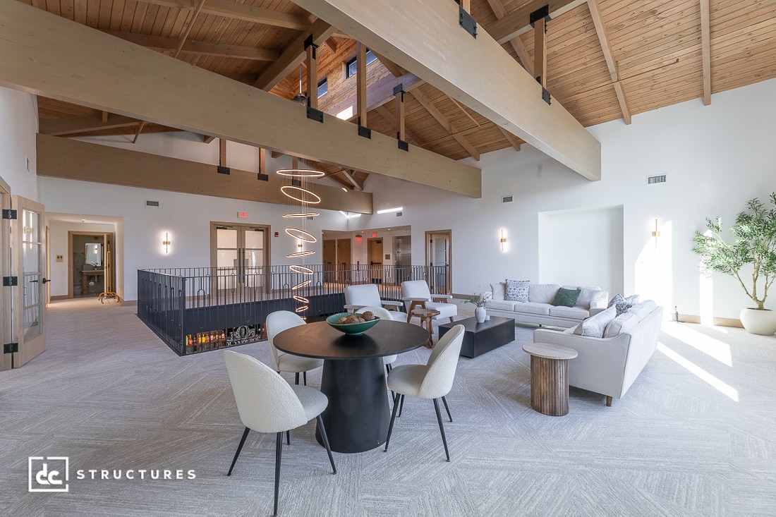 A spacious modern loft with wood-beamed ceilings, neutral furnishings, round dining table, cozy seating area, and large windows.