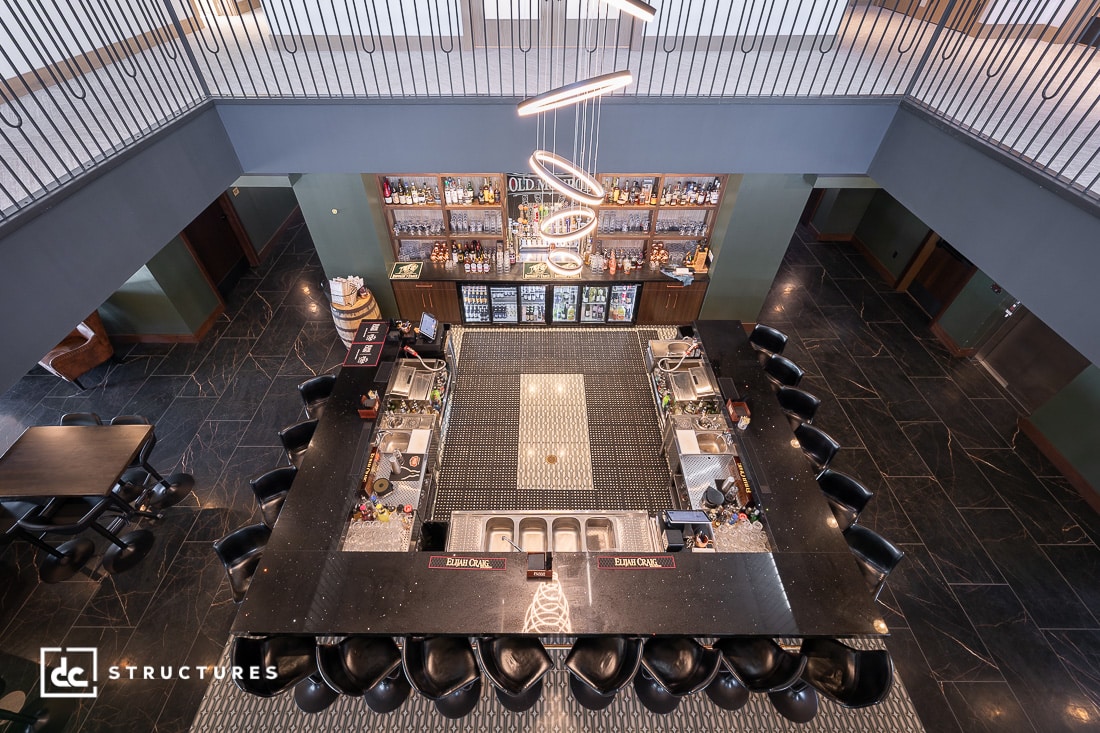 A modern, square-shaped bar with a black marble countertop and black chairs, viewed from above, with shelves, bottles, refrigerators, and a geometric chandelier overhead.