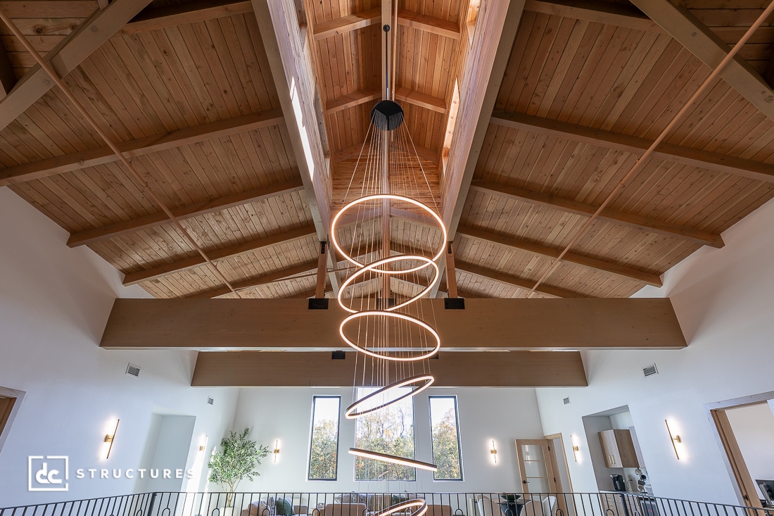 Modern interior with vaulted wooden ceiling, exposed beams, spiral LED chandelier, loft area, tall windows, and minimalist decor.