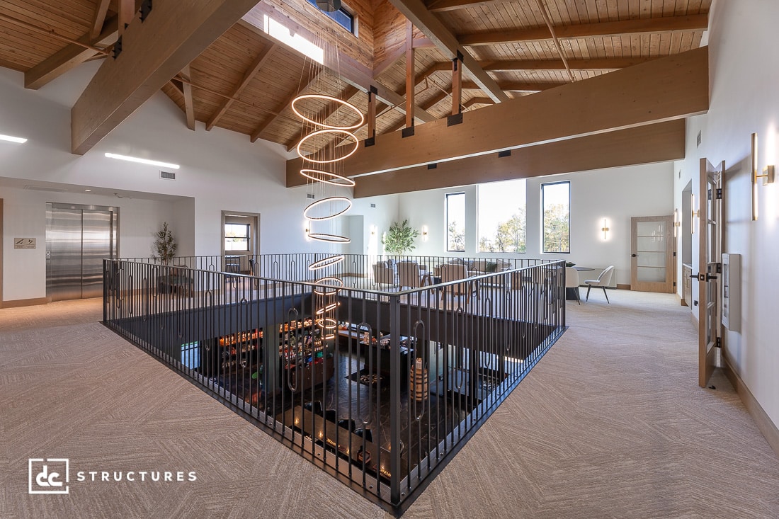 A modern open loft with exposed wooden beams, large windows, a spiral light above a black railing, and neutral-toned furniture.