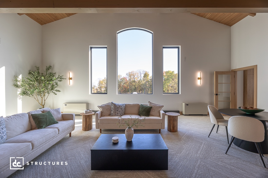 Modern living room with high wood-beamed ceiling, arched center window, side windows, neutral furnishings, and soft natural light.