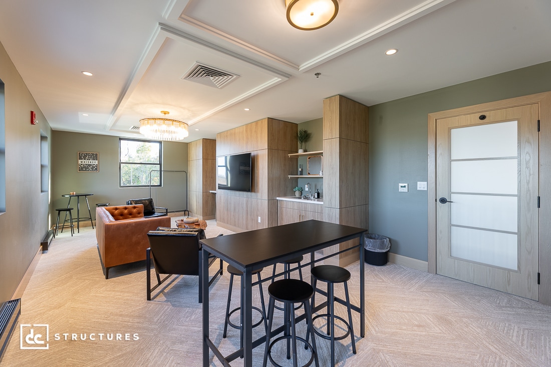 A modern living space with a table and stools in the foreground, lounge area with leather chairs, TV, wood cabinetry, green walls, and large windows letting in natural light.