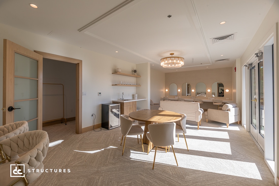 A modern, sunlit lounge with a round table and four chairs, small kitchenette, mirrors, seating area, and large bright windows.