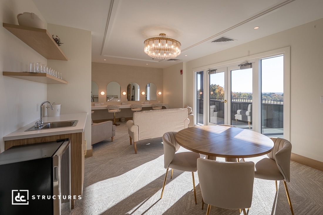 A modern room with a round dining table, four chairs, a small kitchenette, sofas, vanity stations with mirrors, and glass doors to a scenic balcony.