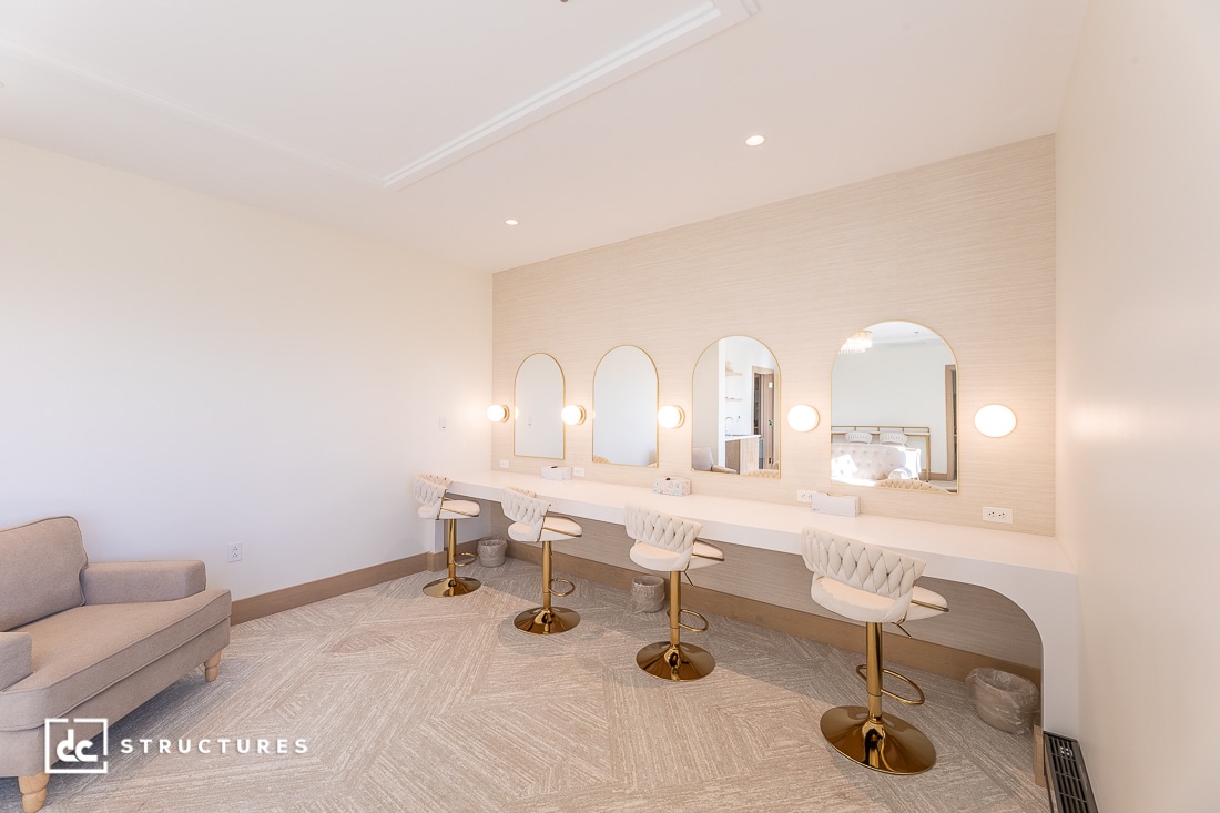 A modern, well-lit vanity room with four gold and white chairs at a long counter, mirrors with built-in lights, and neutral tones.