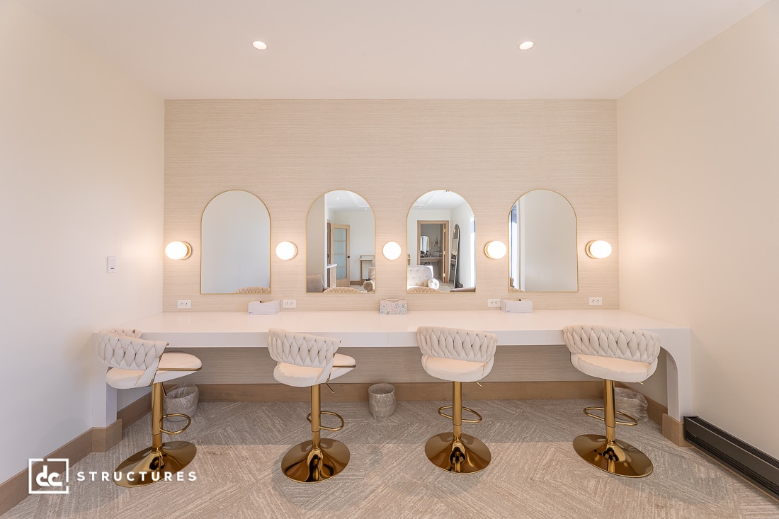 A modern vanity area with four gold and white cushioned chairs, four gold-framed mirrors, and wall sconces in a bright room.