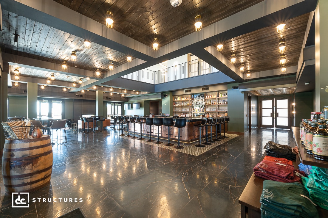 A spacious, modern bar with a marble floor, central bar seating, pendant lights, wood-paneled ceiling, and stocked shelves.