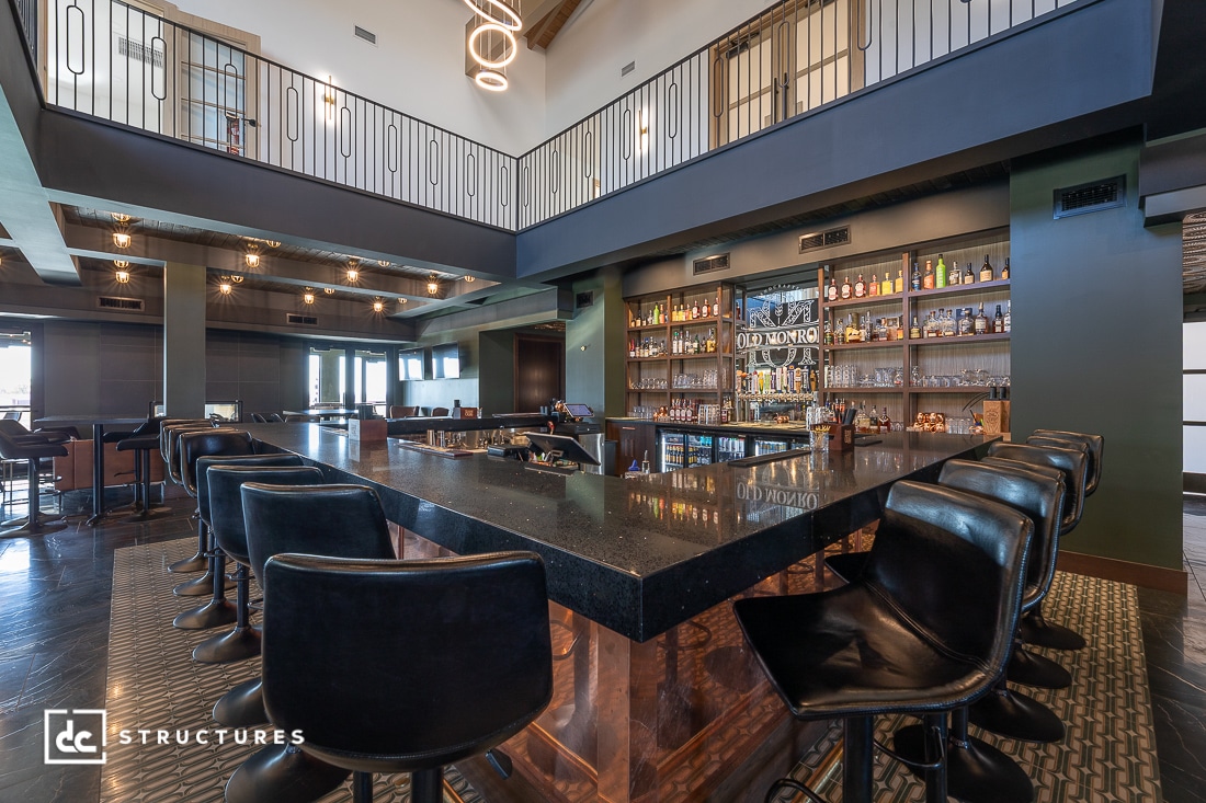 Modern bar interior with black countertops, high-backed swivel chairs, and liquor display. High ceilings and open loft above.