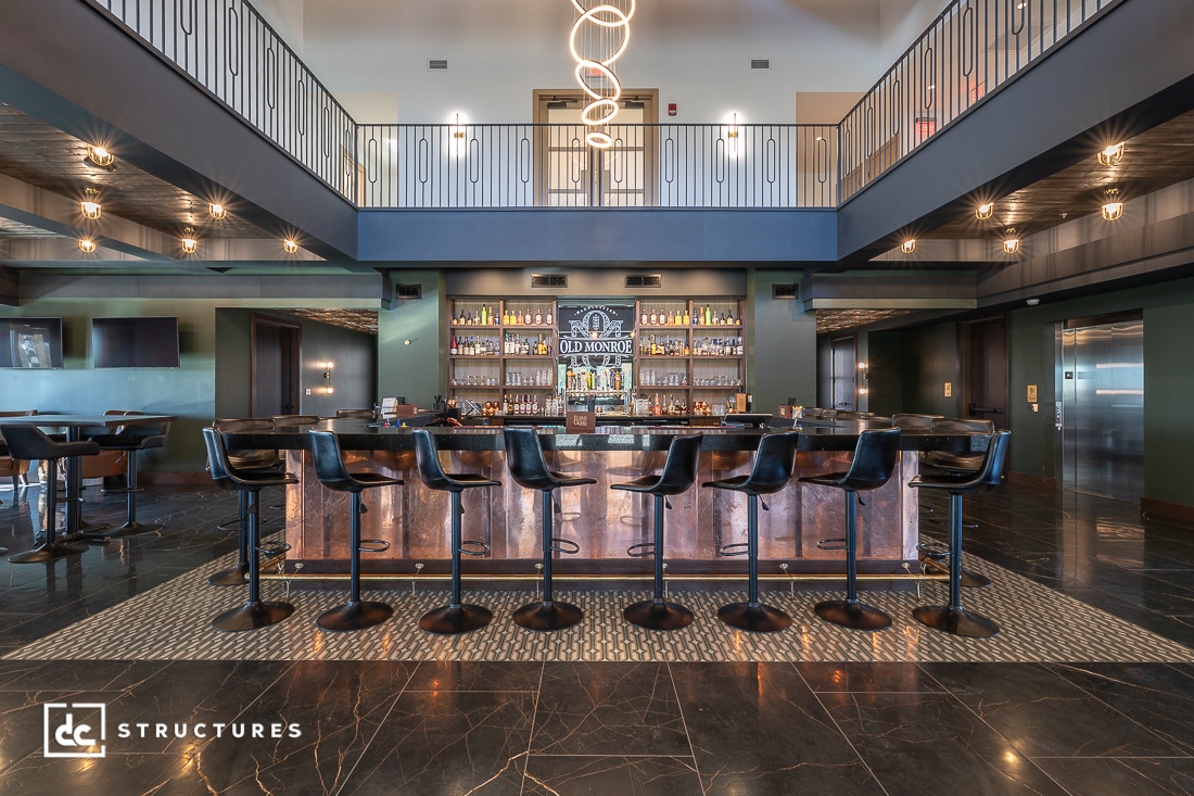 Modern bar interior with a long counter, black barstools, high ceilings, a second-floor balcony, and contemporary lighting fixtures.