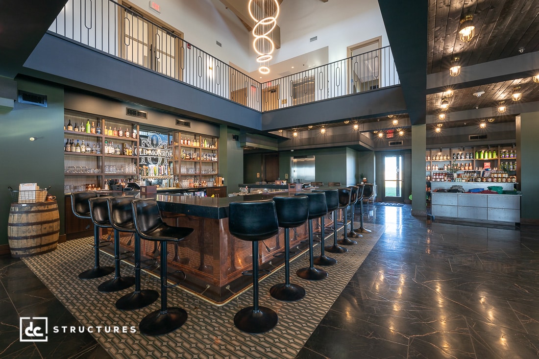 A modern, spacious bar with a central U-shaped counter, high black chairs, shelves of bottles, and an open mezzanine above. Warm lighting creates an inviting atmosphere.