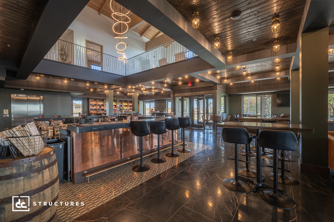 Modern bar and restaurant interior with dark wood ceiling, hanging lights, high tables, bar stools, central bar, and a visible mezzanine.