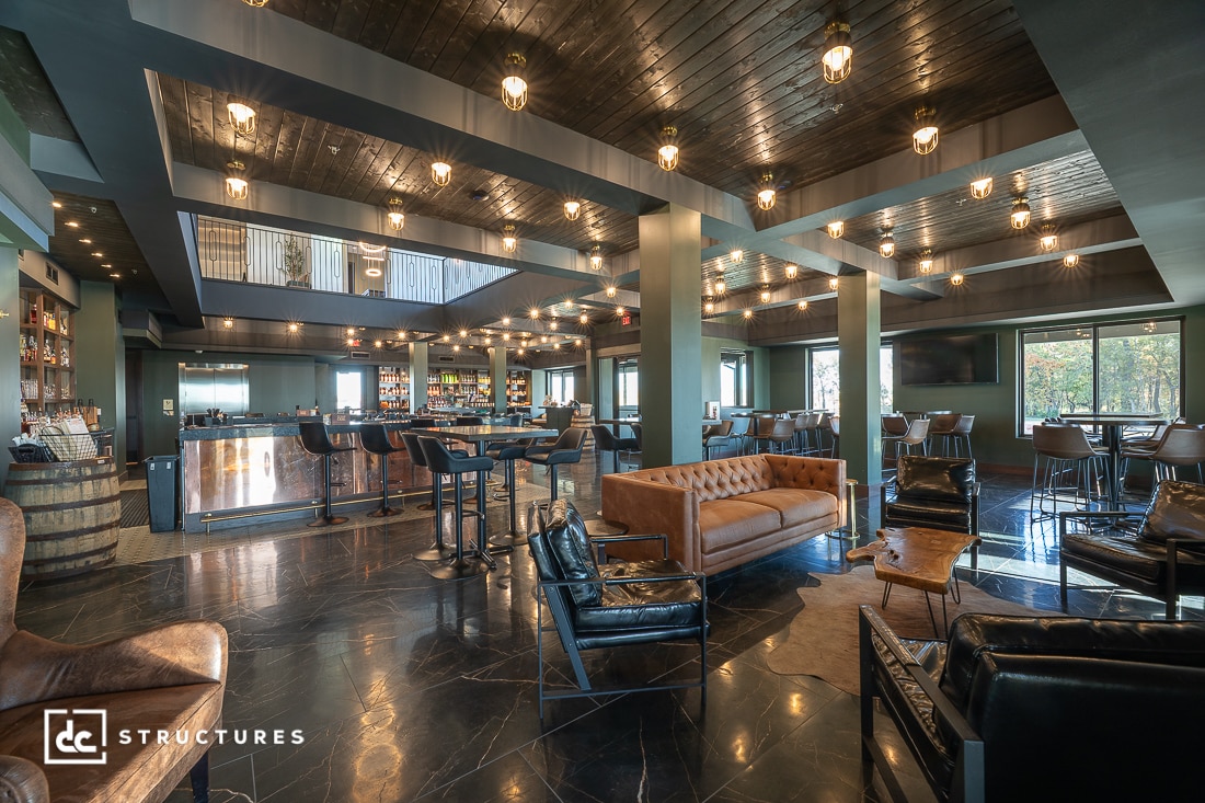 A modern lounge with leather sofas, armchairs, high tables, a bar area, and large windows. Exposed bulbs hang from a dark wood ceiling. Polished black floors reflect the stylish open space.