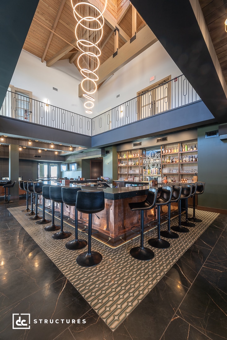 Modern bar area with black swivel chairs around a central bar, backlit shelves of bottles, high ceilings, and circular chandeliers.