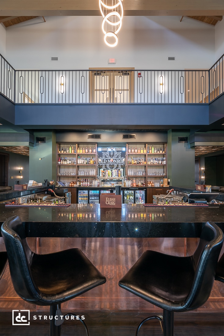 A modern bar interior with two black chairs at a glossy counter, liquor bottles on shelves, mezzanine above, and circular chandelier overhead.