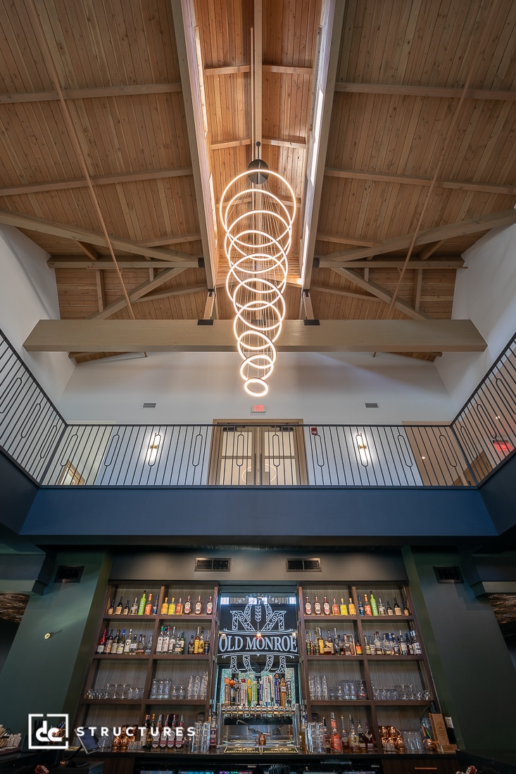 A modern bar interior with a high wooden ceiling, spiral pendant light, and balcony railings. The shelves are stocked with bottles.