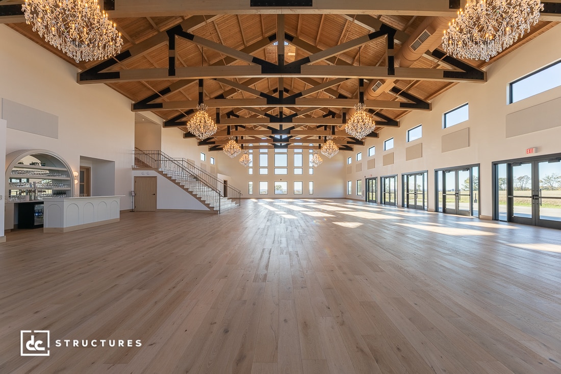 A spacious, elegant hall with high wooden beams, large windows letting in natural light, crystal chandeliers, light wood flooring, and a bar area near a staircase.