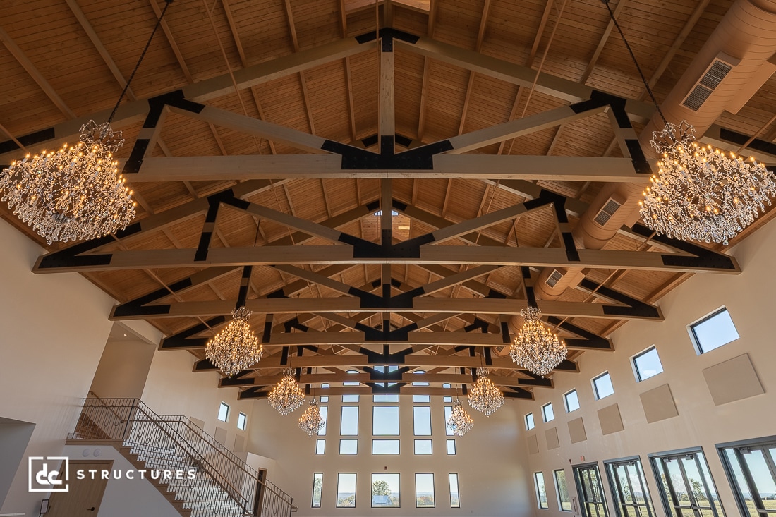 Spacious hall with exposed wooden beams, elegant chandeliers, large windows with natural light, and a staircase on the left.