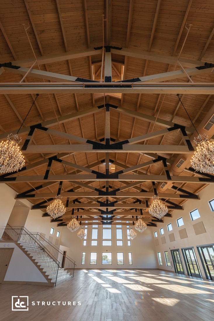 A spacious, sunlit hall with high wooden beam ceilings, large windows, elegant chandeliers, and a staircase along the left wall. The room features light wood floors and modern, clean architectural details.