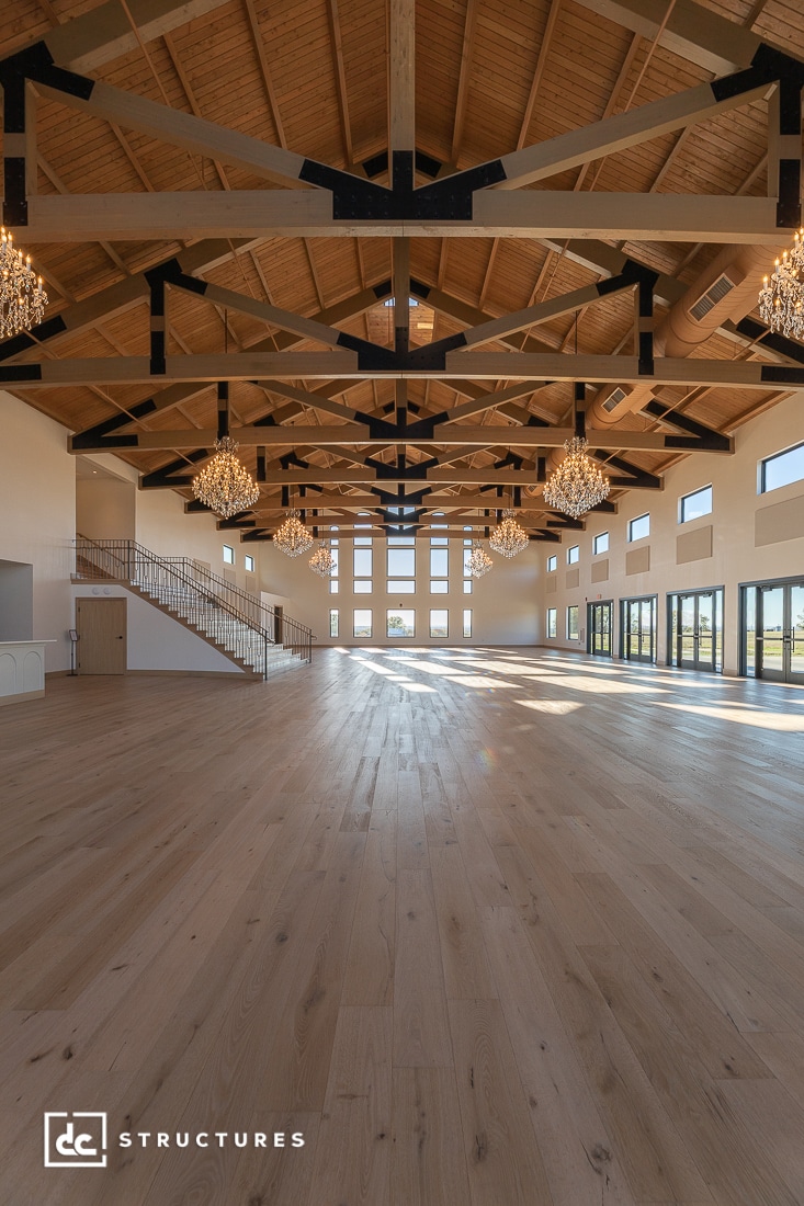 Spacious, empty event hall with high wooden ceilings, exposed beams, and large windows. Chandeliers hang above light wood floors.