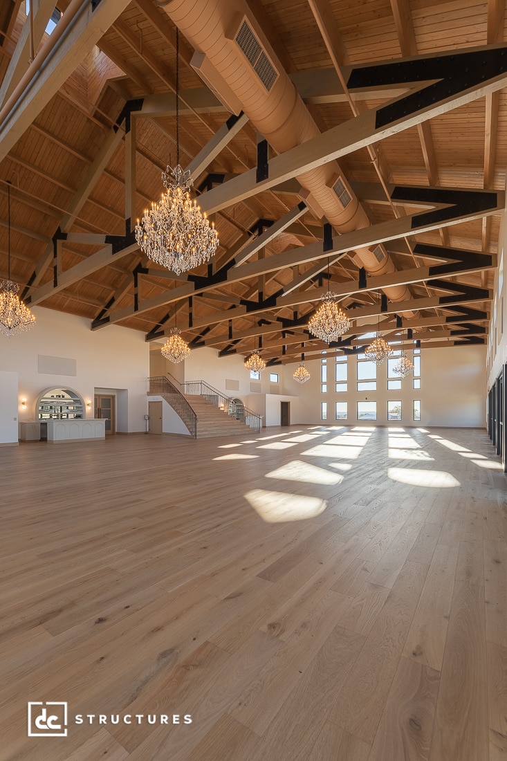 Spacious hall with high wooden ceilings, large windows, chandeliers, light wood floor, staircase, and bar area in natural sunlight.