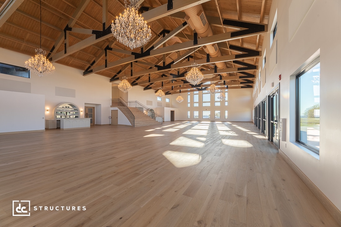 A spacious, sunlit event hall with wooden floors, high vaulted ceilings, exposed beams, large windows, chandeliers, and a staircase.