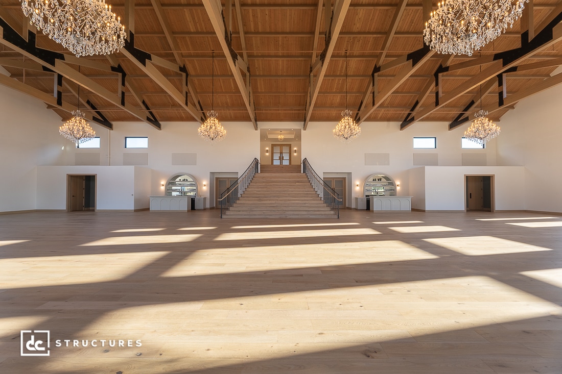 Spacious, elegant hall with high wooden ceiling, chandeliers, wide central staircase, large windows, and light wood flooring.