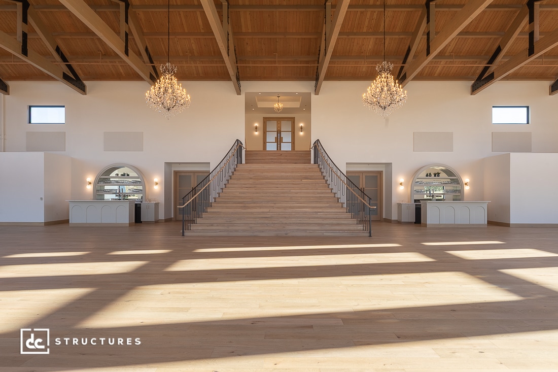 Spacious, elegant hall with a grand central staircase, high wood-beam ceiling, three chandeliers, arched windows, double doors.