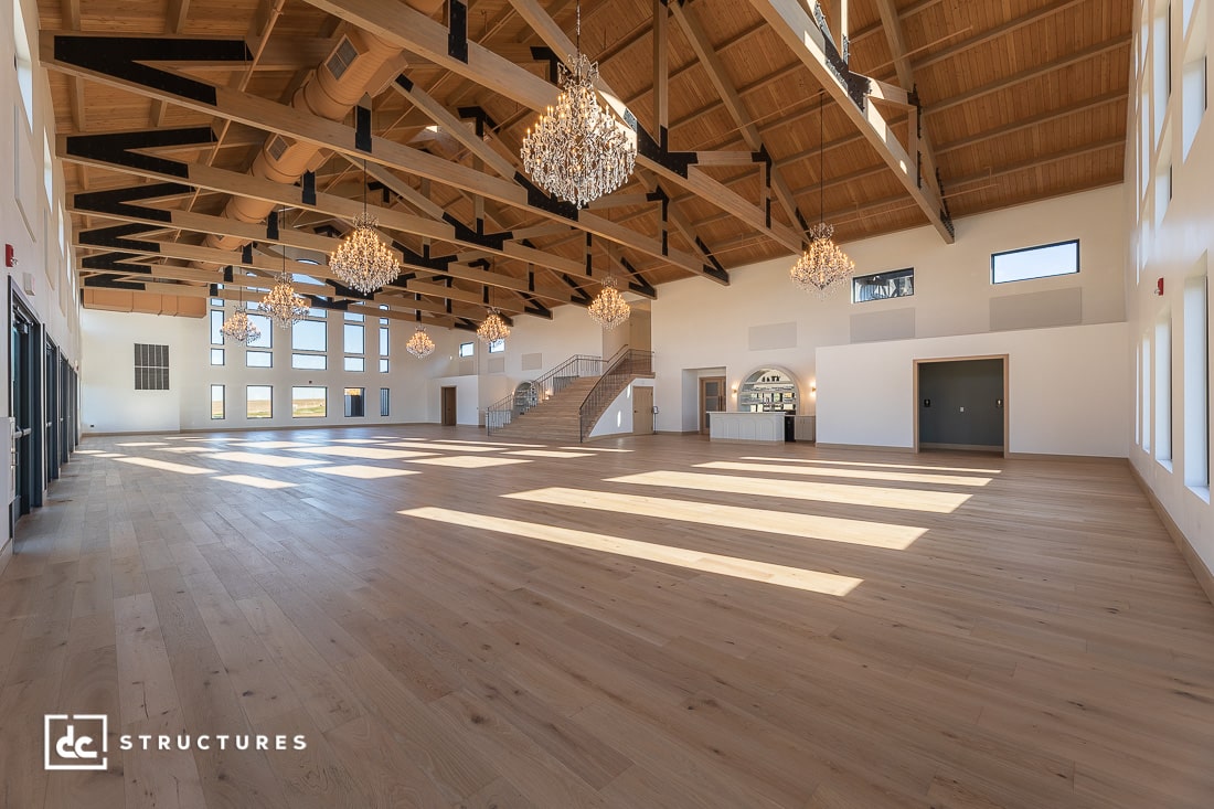 Spacious event hall with high wooden-beam ceilings, large windows, chandeliers, light wood floors, and a central staircase.