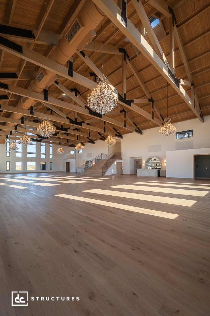 A spacious, elegant hall with wooden floors, high vaulted ceilings, exposed beams, large windows, chandeliers, and a staircase.