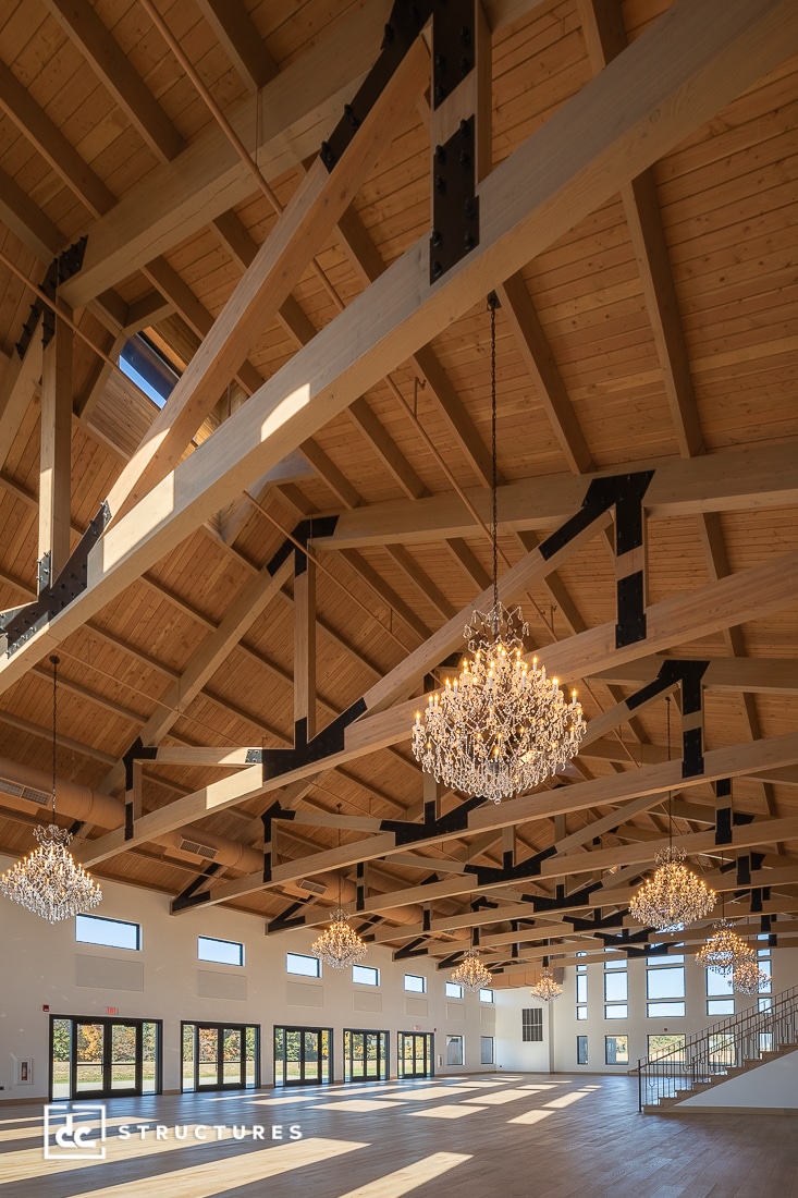 Spacious event hall with high wooden beam ceiling, large windows, and crystal chandeliers. Sunlight highlights the airy atmosphere.