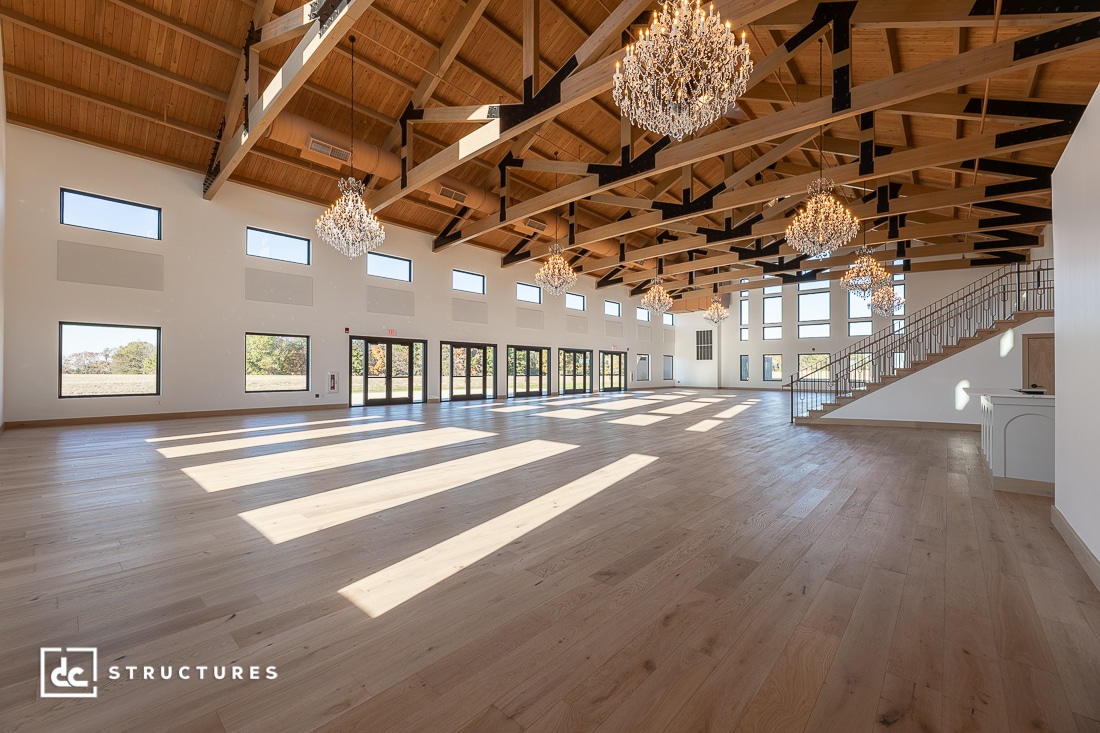 Spacious sunlit hall with high wooden ceilings, exposed beams, large windows, elegant chandeliers, and light hardwood floors.