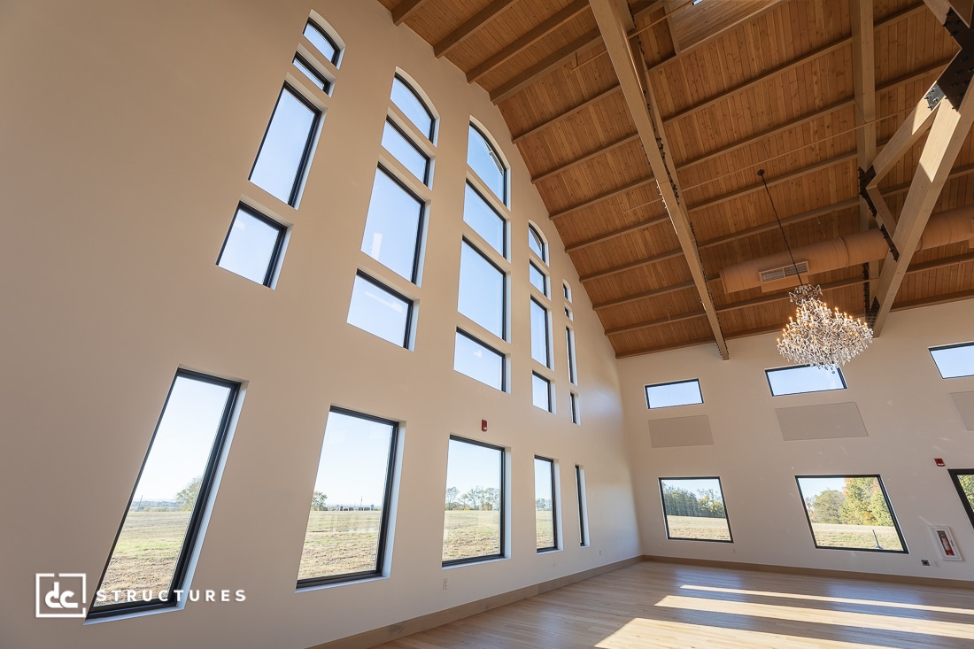 A spacious room with high vaulted ceilings, large arched and rectangular windows, wood beams, a chandelier, and light-colored walls and floors.