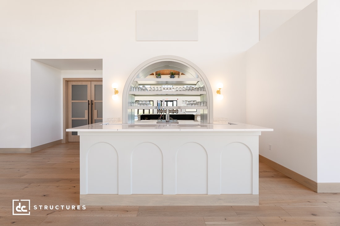 A modern, minimalist bar with a white countertop, arched accents, and a large arched mirror behind it. Light wood floors and white walls with ample natural light.