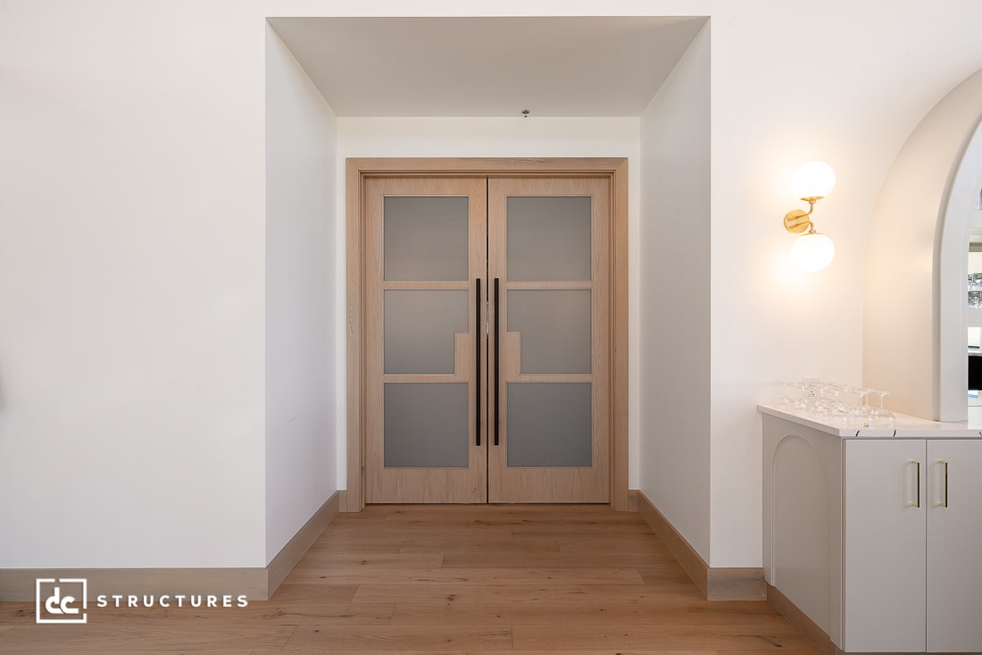 Double wooden doors with frosted glass panels in a white hallway with light wood flooring and a modern wall sconce above a counter.