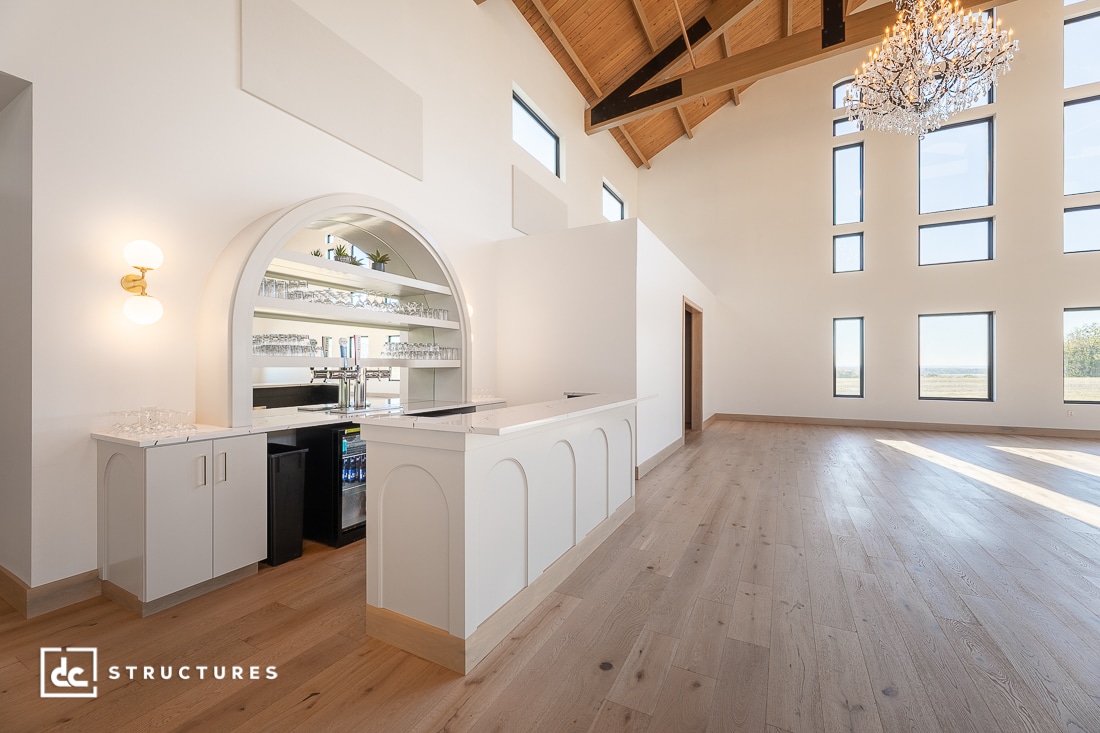 Modern, spacious event hall with high ceilings, tall windows, wooden floors, a chandelier, and a built-in bar. Natural light fills the airy room.