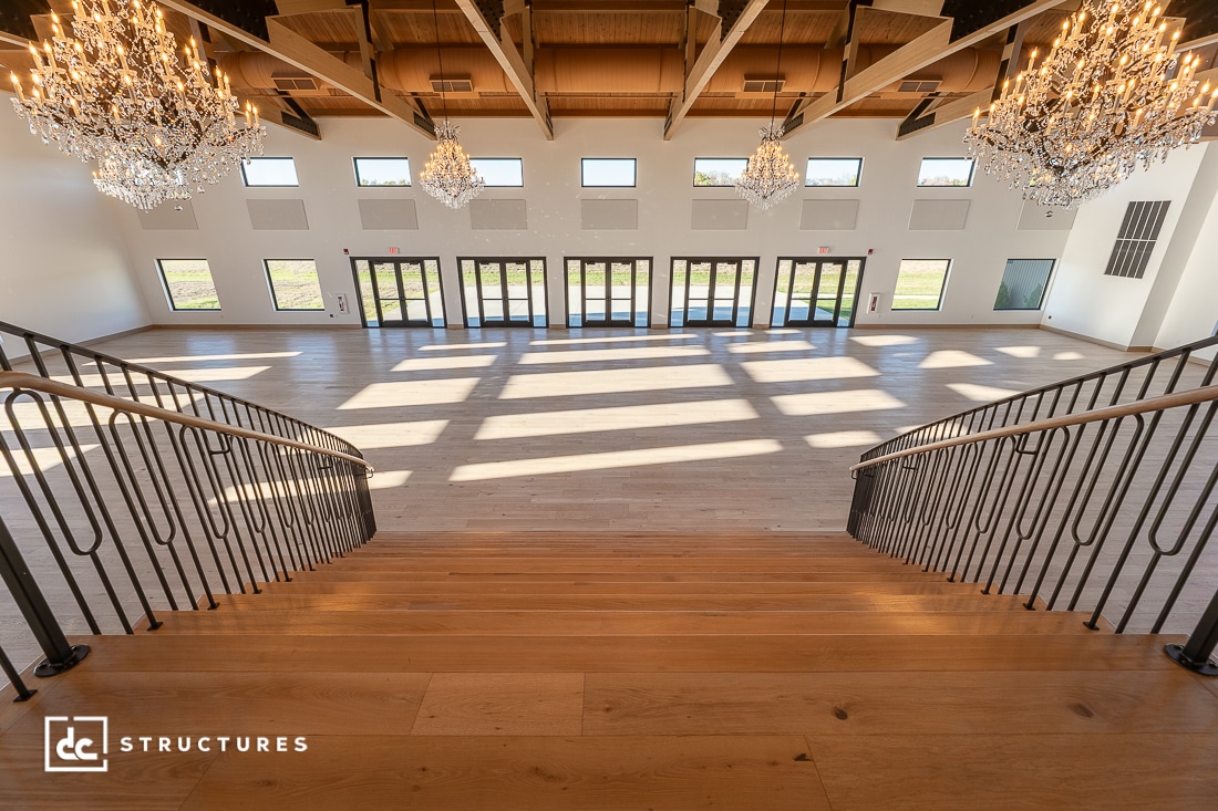 A spacious, bright event hall with wooden floors, large windows, chandeliers above, and a wide staircase with metal railings.