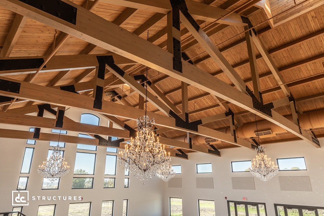 The image shows a spacious room with high ceilings, exposed wooden beams, large windows, and elegant crystal chandeliers. Sunlight streams in.