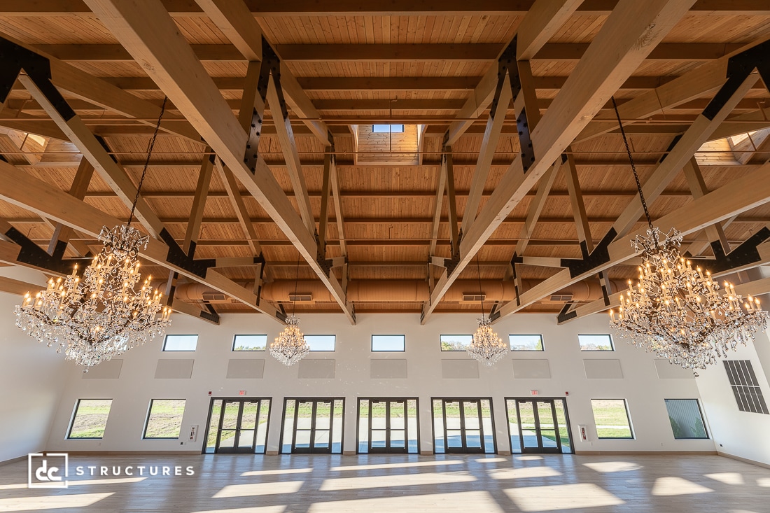 A spacious hall with exposed wooden beams, large windows, glass doors, and hanging crystal chandeliers. Sunlight streams in.