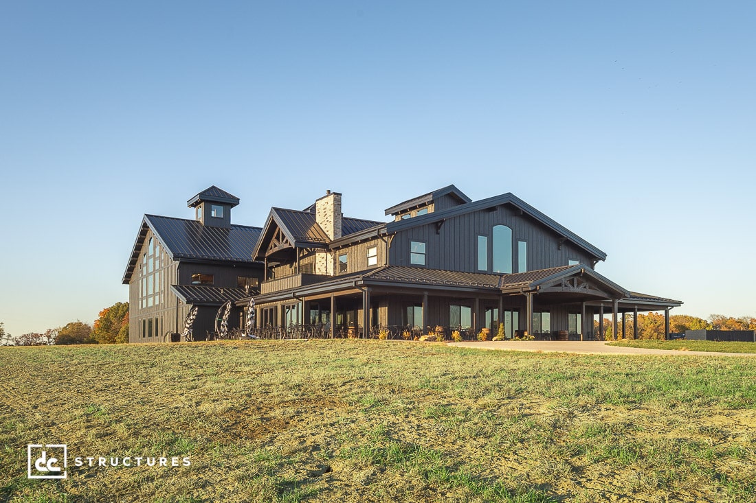 A large modern barn-style house with dark siding and a metal roof sits in a grassy field under a blue sky, with multiple rooflines and a cupola tower.