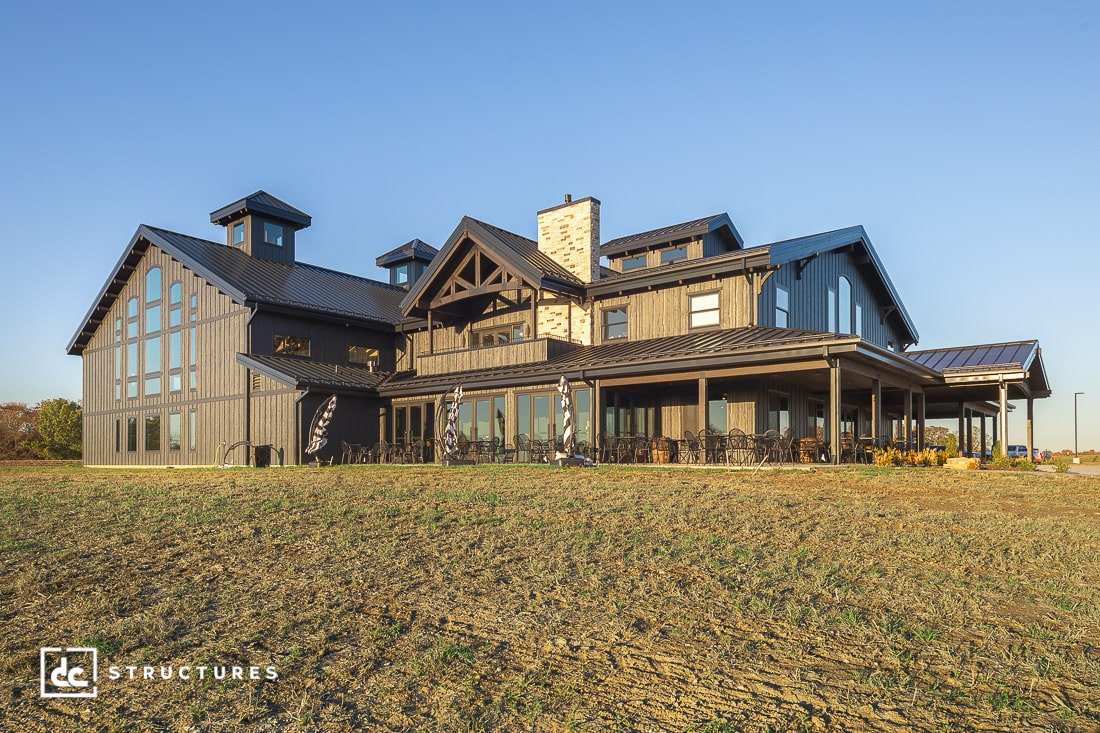 A large modern farmhouse with tall windows, gray siding, and black metal roof sits on a grassy plot under a clear blue sky.