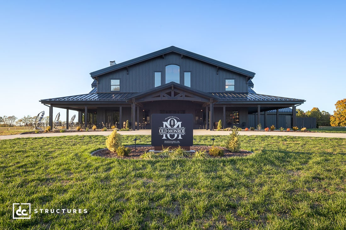 A large, dark gray barn-style building with a wraparound porch is surrounded by grass, landscaping, and a blue sky.