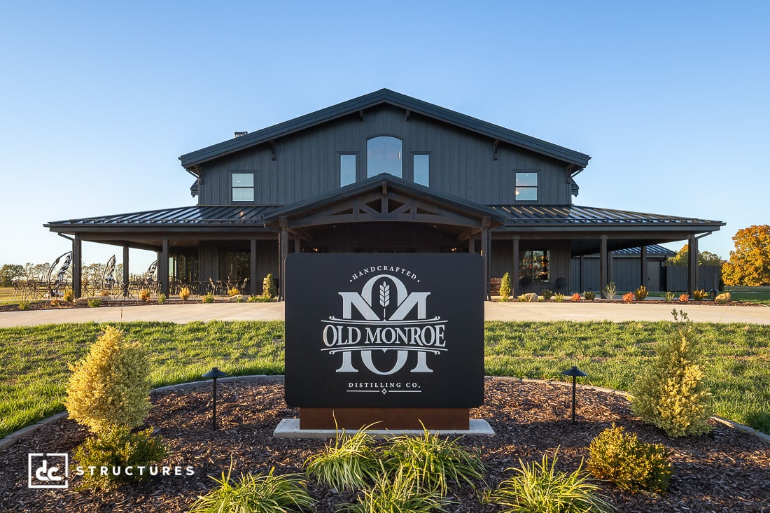 A large, modern barn-style building with dark exterior siding, a gabled roof, and covered porches sits behind a landscaped area.