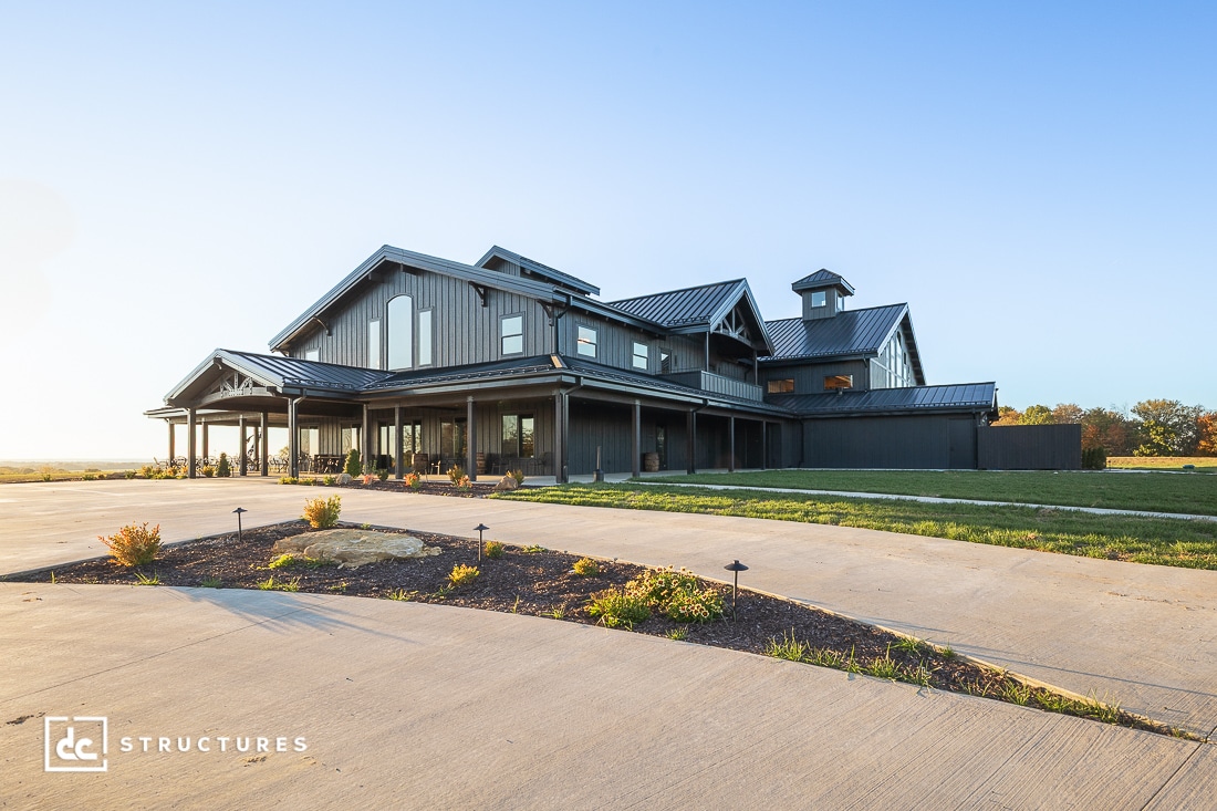 A large modern farmhouse-style building with black siding, multiple gables, and a cupola sits on a spacious lot at sunset.