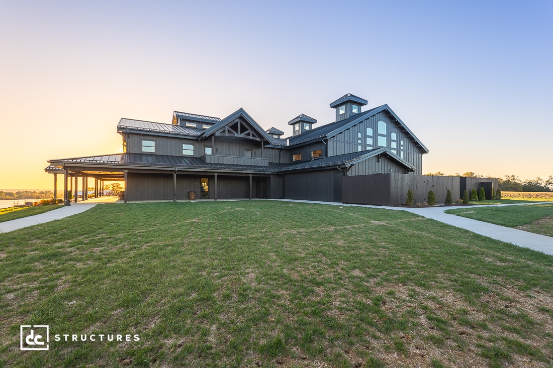 Large modern barn-style building with dark wood siding, multiple peaked roofs, and cupolas, surrounded by green grass and a concrete walkway at sunset.