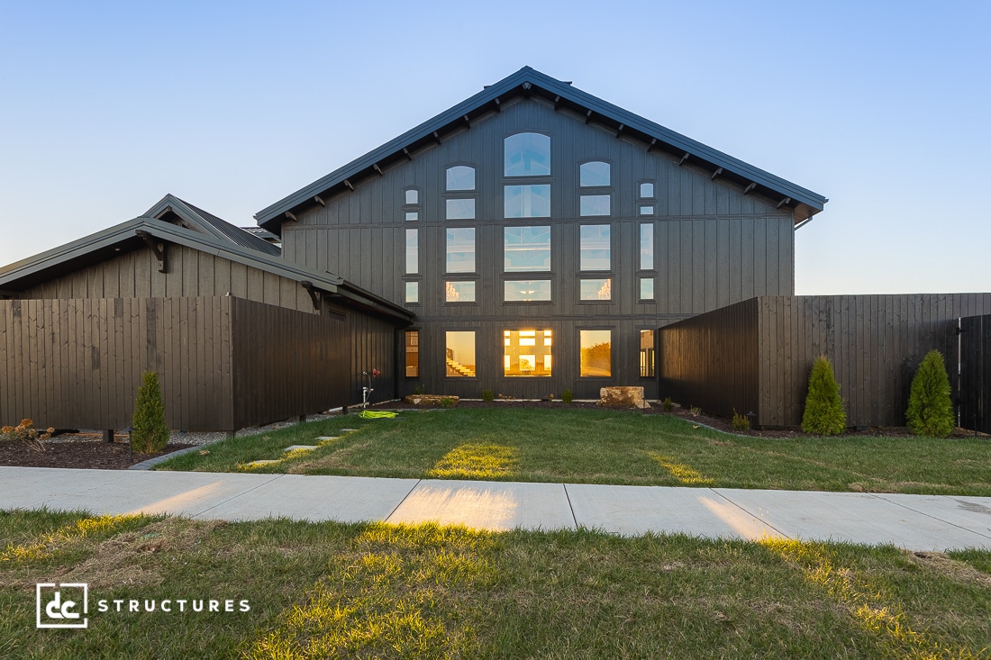 A modern barn-style building with large windows reflecting the sunset, surrounded by black fencing, green grass, and small shrubs.
