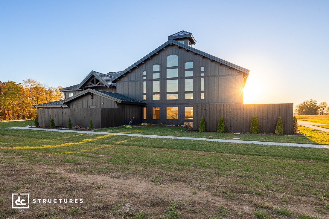 A modern, dark wood barn-style building with large windows stands on a green lawn at sunset, surrounded by trees and open fields under a clear sky.