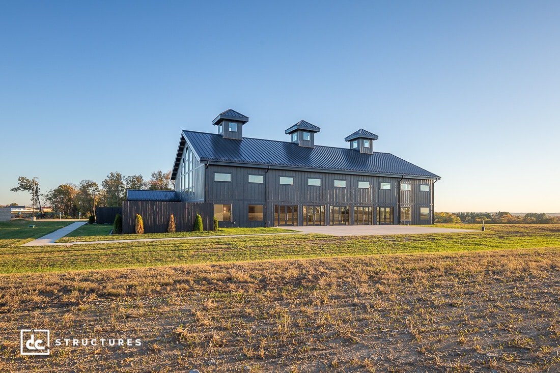 Modern barn-style building with large windows, dark exterior, and three cupolas on the roof, on a grassy field under blue sky.