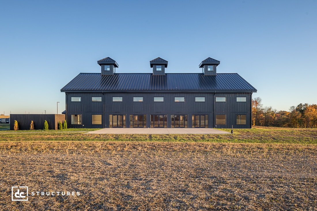 A large, modern black barn with three cupolas sits on a flat, grassy area under a clear blue sky. The structure features multiple windows and garage-style doors.