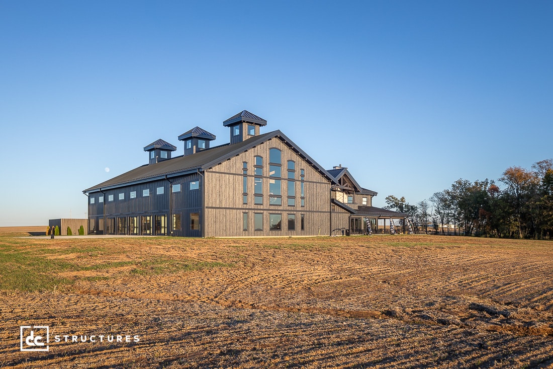 A large modern barn-style building with three cupolas sits on a flat open field under a clear blue sky, with trees in the background.